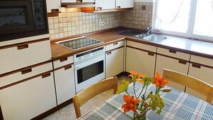 A compact kitchen with white cabinets, a microwave, a dishwasher, and a sink. There's a window with curtains and a table with a plaid tablecloth and flowers. - Vacation home Roth by Interhome (Ichenheim)