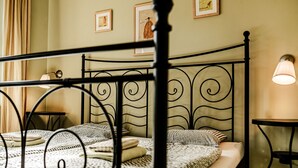 Ein Bett mit Metallgestell, ein Nachttisch und eine Lampe. An der Wand hängen Bilder in Rahmen.