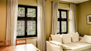 Ein Zimmer mit einem Holztisch und Stühlen, einer Couch mit Kissen, zwei Fenstern mit schwarzen Rahmen und weißen Vorhängen sowie einer Pendelleuchte.