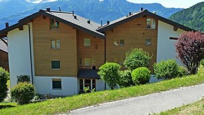 A two-story wooden building with a sloped roof, surrounded by greenery and mountains in the background. - Seya 19 by Interhome (Ovronnaz)