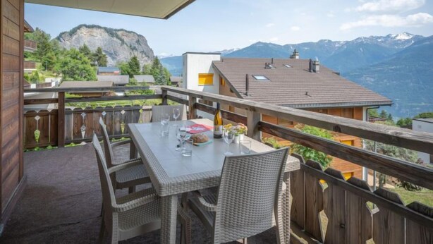 A balcony with a table and chairs, overlooking a mountainous landscape with a view of a lake and buildings. - Seya 19 by Interhome (Ovronnaz)