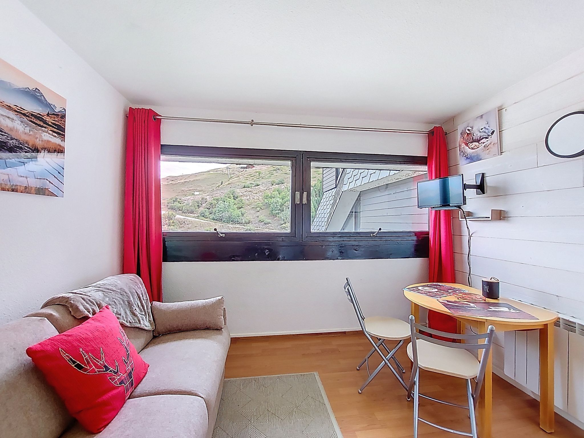 A small, cozy room with a sofa, a small table, a chair, a window with a view of a hilly landscape, and a mounted TV.