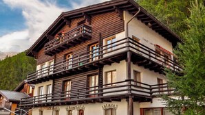 Una cabaña de madera de varios pisos, con balcones, base de piedra y montañas al fondo.