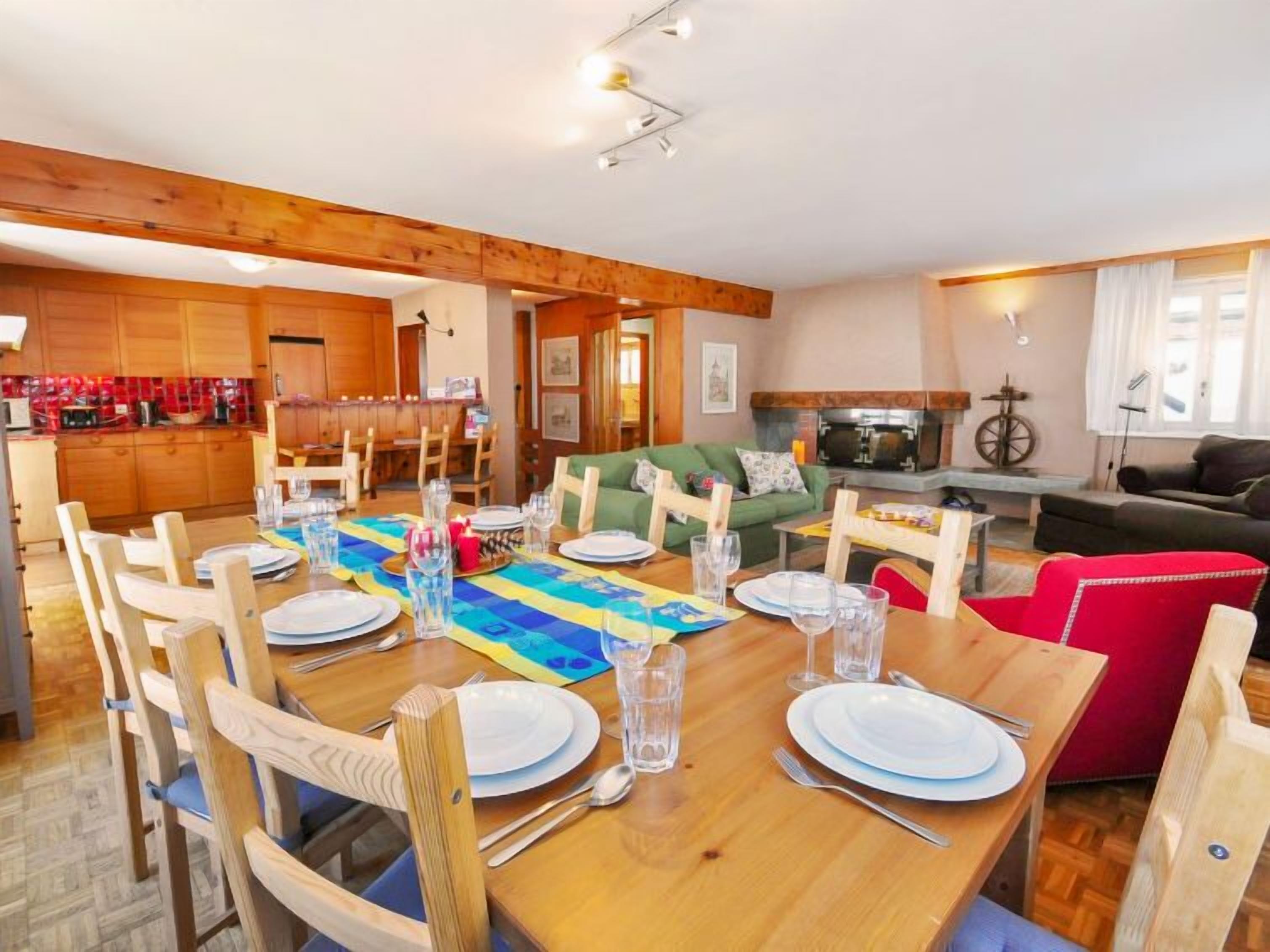 Un coin repas avec une table en bois dressée pour quatre personnes, entourée de chaises en bois. Un canapé vert et un fauteuil rouge sont visibles à l’arrière-plan.