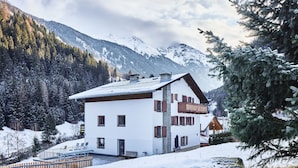 Un chalet bianco con tetto spiovente, persiane in legno e un balcone. È circondato da terreno e alberi innevati, con montagne sullo sfondo.