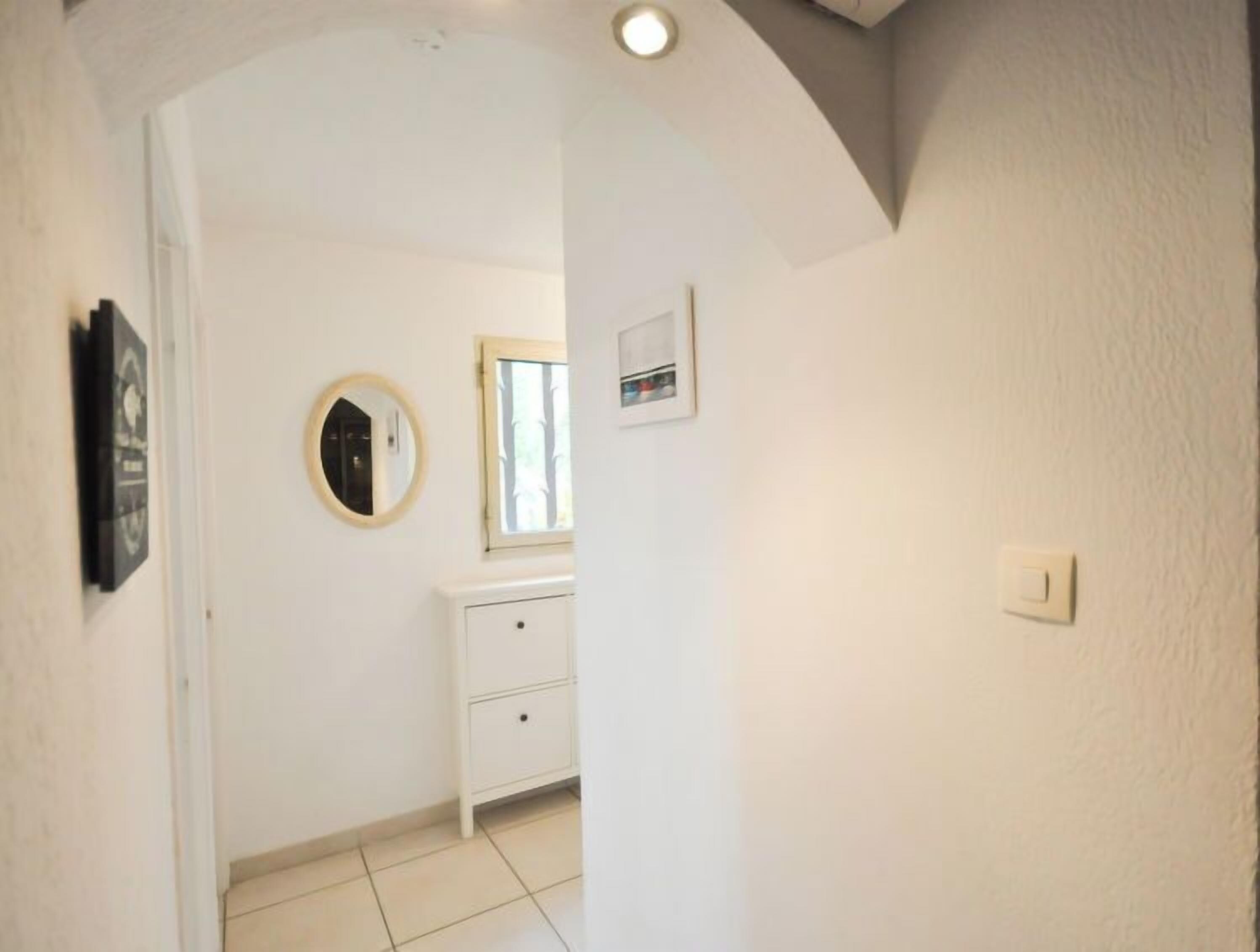 Un couloir étroit avec un mur blanc, un miroir rond, une petite fenêtre et un meuble blanc.