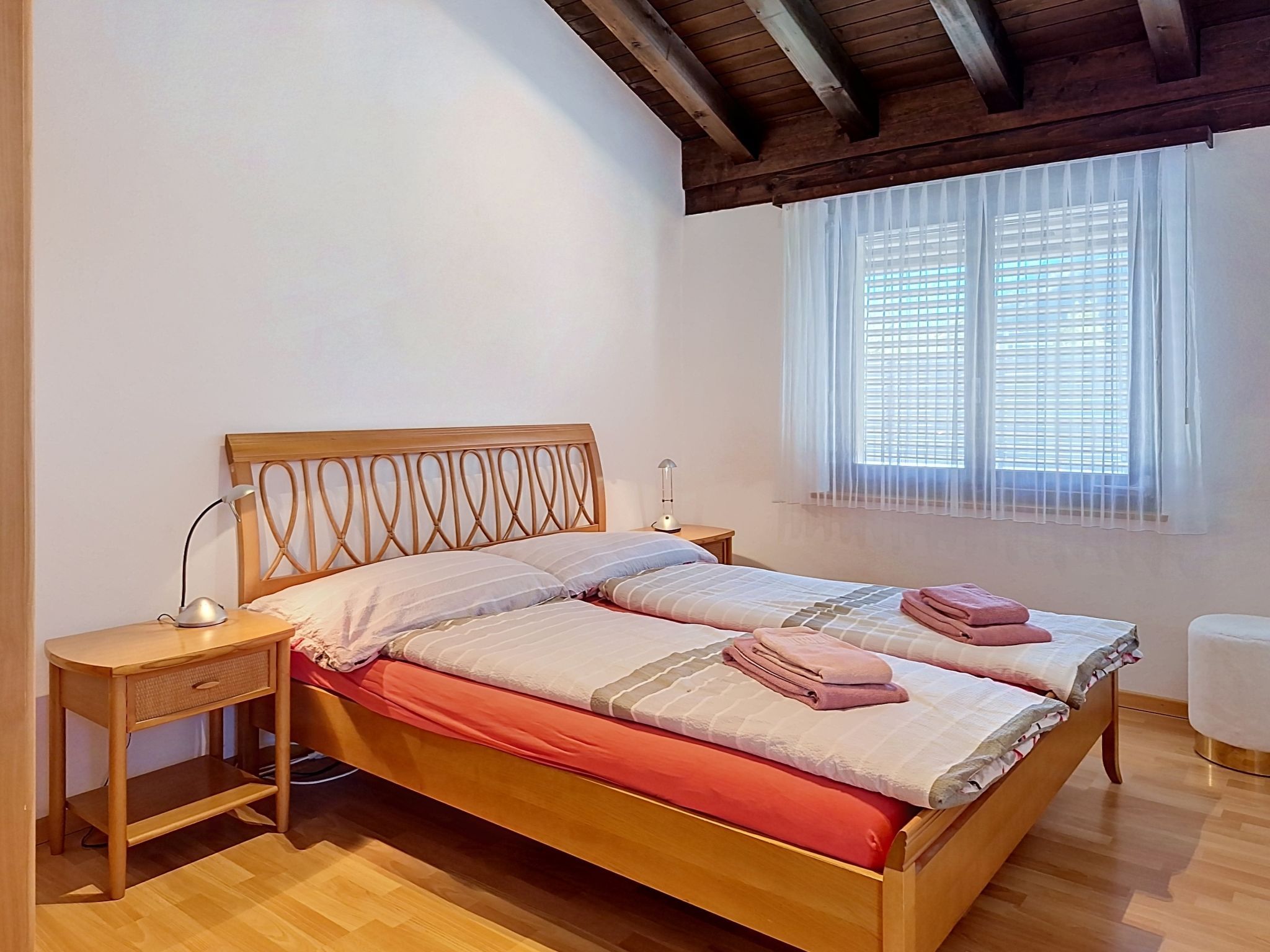 A double bed with a wooden headboard, two bedside tables, a window with blinds, and a wooden floor.