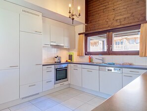 Une cuisine moderne avec des meubles blancs, un four encastré, un lave-vaisselle et une fenêtre avec des rideaux.