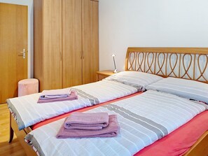 Ein Doppelbett mit gestreifter Bettwäsche und zwei gefalteten Handtüchern, ein Holzschrank, ein Nachttisch mit Lampe sowie ein kleiner Hocker.