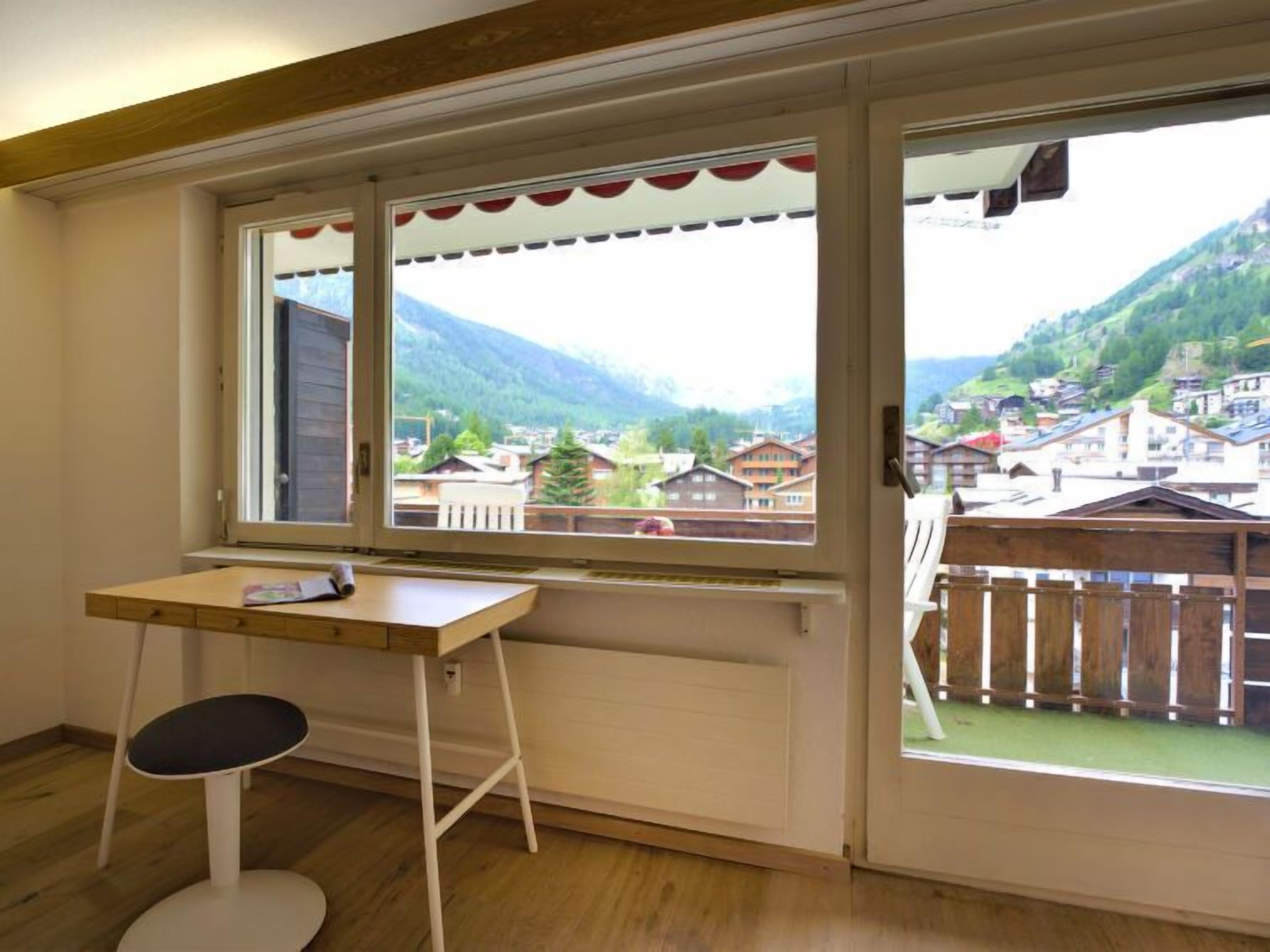 Een kamer met een houten bureau, een stoel en een groot raam met uitzicht op een dorp en bergen.