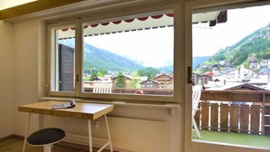 Une pièce avec un bureau en bois, une chaise et une grande fenêtre donnant sur un village et des montagnes.