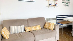 En stue med en beige sofa, mønstrede puder, et indrammet maleri, en hvid reol med dekorative genstande, og et spiseområde med en stribet stol.
