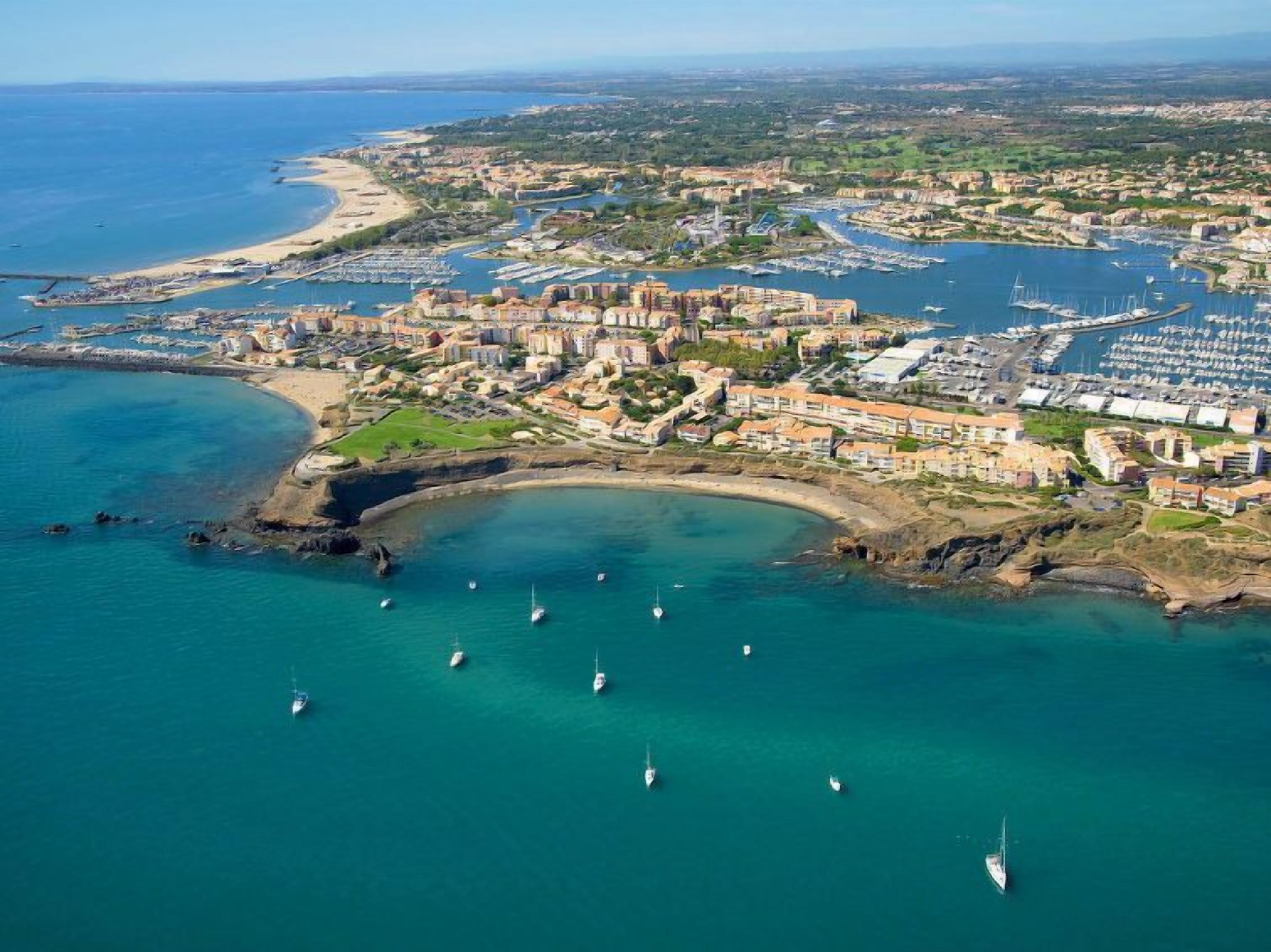 An aerial view of a coastal town with a marina, sandy beaches, and residential buildings.