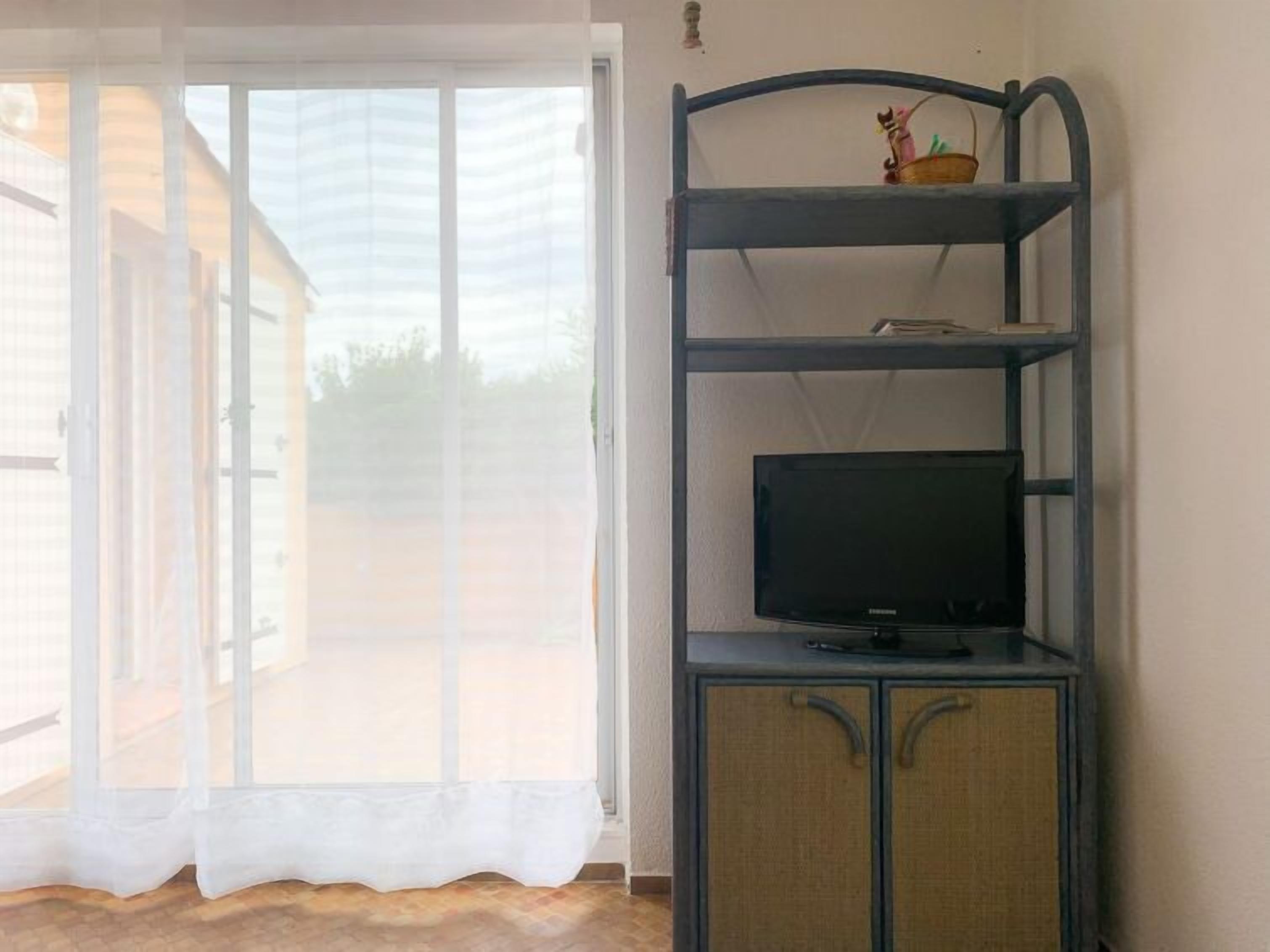 A room with a sliding glass door leading to an outdoor area, a television on a wooden cabinet, and a small shelf with decorative items.