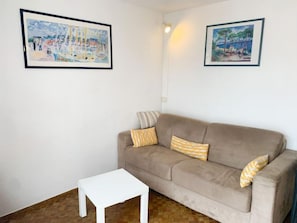 Un salon avec un canapé beige, deux coussins à motifs, une table basse blanche et deux tableaux encadrés accrochés aux murs.