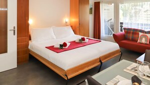 A hotel room with a bed, a red sofa, a dining table set with plates and glasses, and a balcony with a railing. - Matten (Utoring) by Interhome (Zermatt)