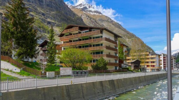 A multi-story hotel with balconies, a river in the foreground, and mountains in the background. - Matten (Utoring) by Interhome (Zermatt)