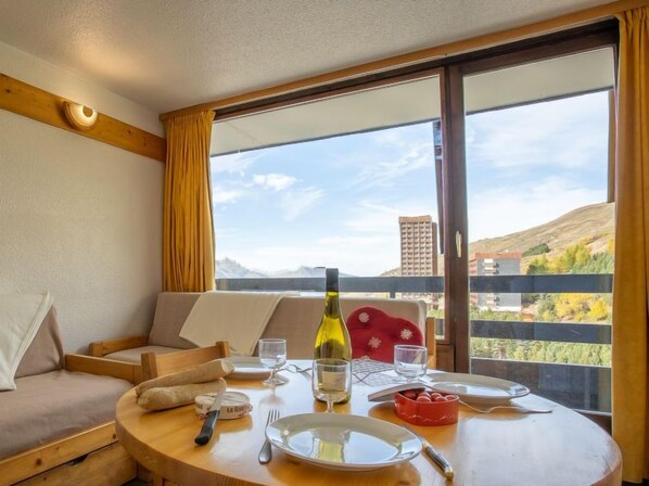 Un coin repas avec une table dressée pour deux personnes, une bouteille de vin et une vue panoramique sur les collines et les bâtiments, visible à travers de grandes fenêtres.