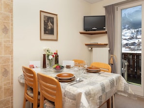 Uma sala de jantar com uma mesa posta para dois, uma televisão acima da mesa e vista para uma paisagem nevada através de uma porta de vidro.