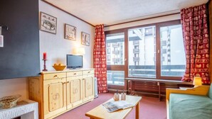Une pièce chaleureuse avec un meuble en bois, une télévision, une petite table sur laquelle repose un journal et une fenêtre ornée de rideaux à motifs.