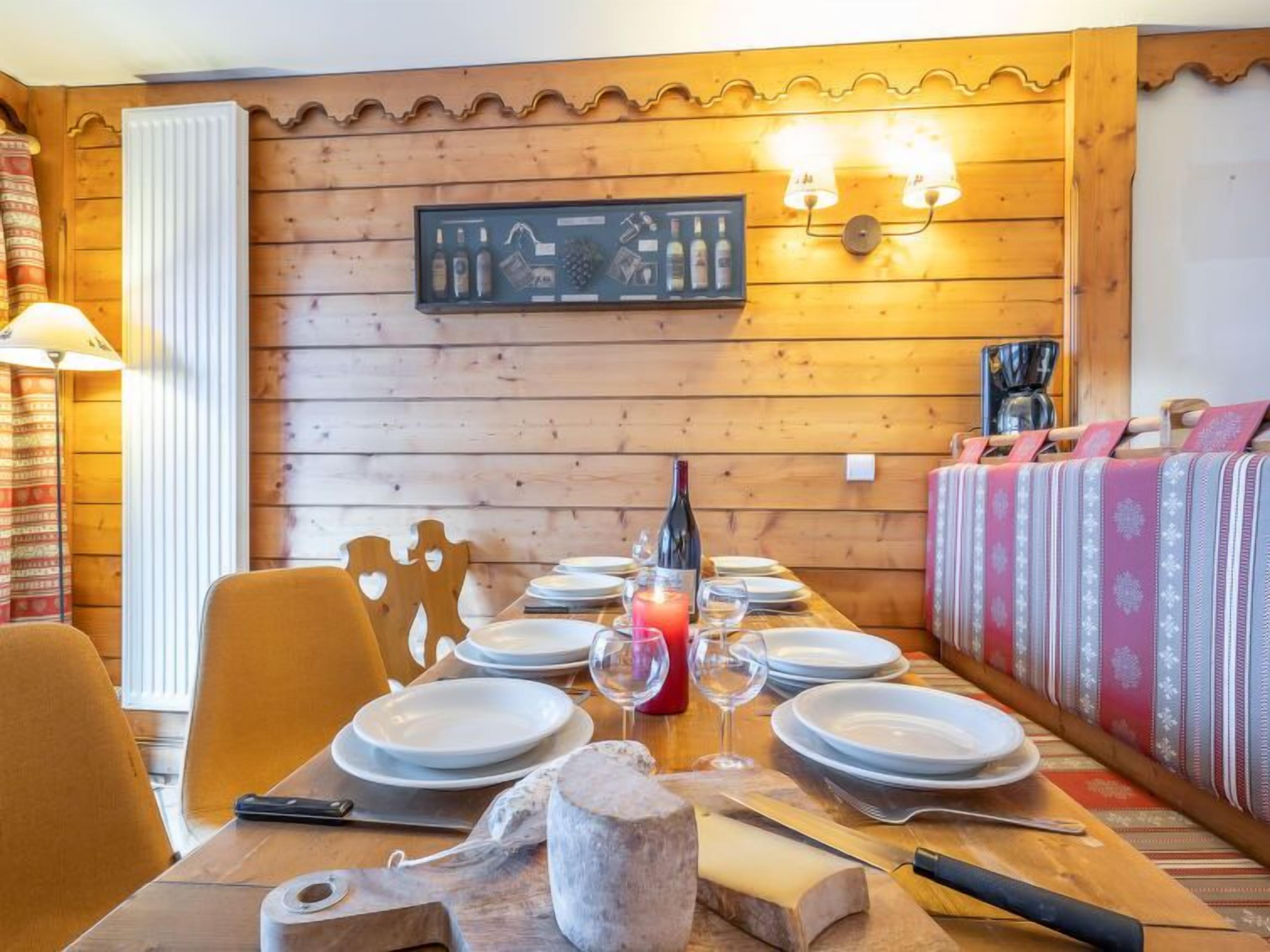 A wooden dining table set with plates, glasses, and a cheese wheel, with a wine bottle and candle on the table.