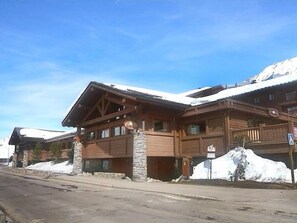 A wooden lodge with stone pillars, a ski slope in the background, and a clear blue sky.