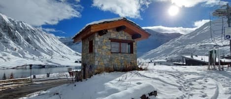 A stone cabin with a wooden roof, surrounded by snow-covered mountains and a clear sky.