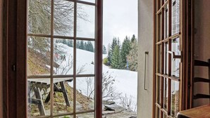 Blick durch eine Glastür auf eine Steinterrasse mit einer hölzernen Bank, eine verschneite Landschaft und ein Wald im Hintergrund.
