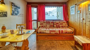 A cozy room with a wooden bench, a table with a bottle, and a window with red curtains.