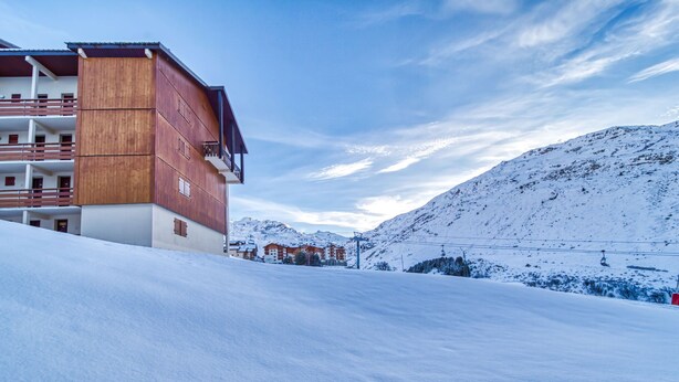 A snow-covered mountain slope with a wooden building featuring balconies and a ski lift in the background.