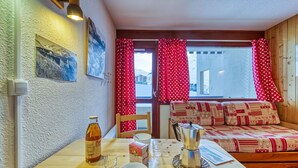 A cozy room with a wooden table, a sofa with patterned cushions, a window with red heart-patterned curtains, and a painting on the wall.