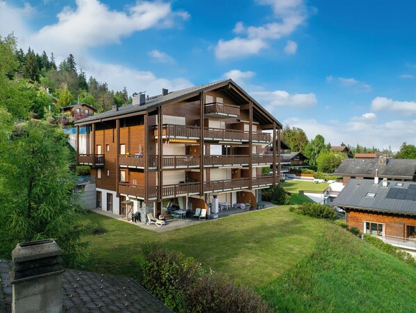 A multi-story wooden building with balconies, surrounded by greenery and a clear sky. - Fénestral R01 by Interhome (Ovronnaz)