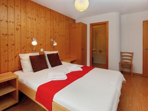 A wooden-paneled bedroom with a bed, two bedside lamps, a red blanket, and a wooden chair. - Fénestral R01 by Interhome (Ovronnaz)