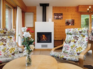 Ein gemütlicher Raum mit Kamin, Holzwänden, floralen Sesseln und einem Blumenstrauß auf einem Tisch.