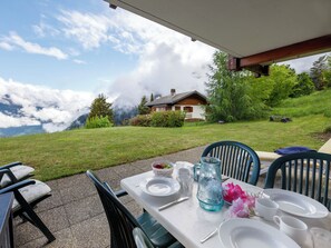 An outdoor dining area with a table set for two, a view of a mountain, and a house in the background. - Fénestral R01 by Interhome (Ovronnaz)