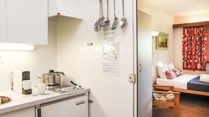Eine kleine Küche mit Kaffeemaschine, Wasserkocher und Herd. Ein kleiner Kühlschrank und ein Spülbecken sind ebenfalls vorhanden. Der Raum verfügt über ein Bett mit Holz Kopfteil und einem roten Vorhang.