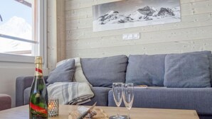 Een gezellige zithoek met een grijze bank, een houten tafel met champagne, twee glazen en een snijplank met een mes.