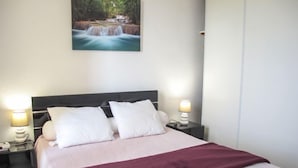Un letto rifatto con cuscini bianchi e una coperta viola, una lampada da comodino, un libro e un quadro raffigurante una cascata sulla parete.