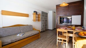 Una habitación acogedora con un sofá de madera, una mesa de comedor con sillas, un armario de madera y una ventana con persianas.