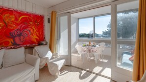 Ein Zimmer mit einem weißen Sofa, einem Tisch mit Stühlen, einem großen Fenster mit Blick auf Bäume und ein Gewässer, sowie einem farbenfrohen, abstrakten Gemälde an der Wand.