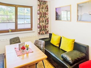 Une pièce comprenant une table, des chaises, un canapé avec des coussins jaunes, un radiateur et une fenêtre avec des stores.