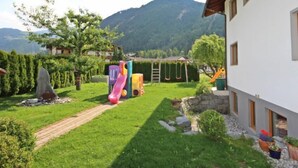 Une maison avec un jardin, une terrasse en bois, un toboggan et une balançoire.