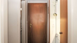 Un couloir étroit, avec une porte en bois au fond, un mur blanc orné d’un tableau d’affichage et une porte blanche sur la droite.