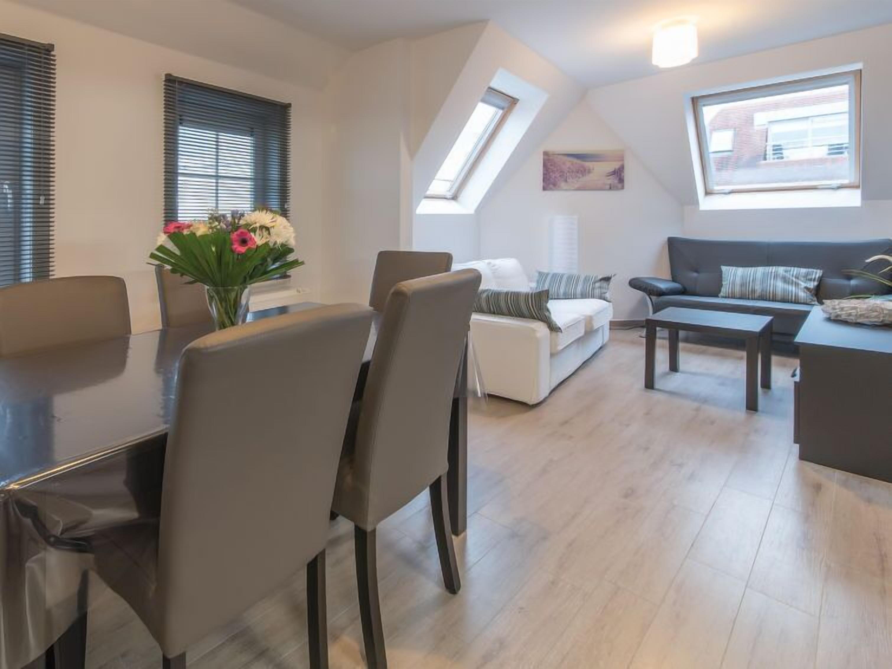 Una sala de estar moderna con comedor, que incluye un sofá, una mesa de centro y una mesa de comedor con sillas. Hay tragaluces y un cuadro en la pared.