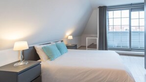 A modern bedroom with a large bed, two bedside tables with lamps, a crib, and a large window with curtains.