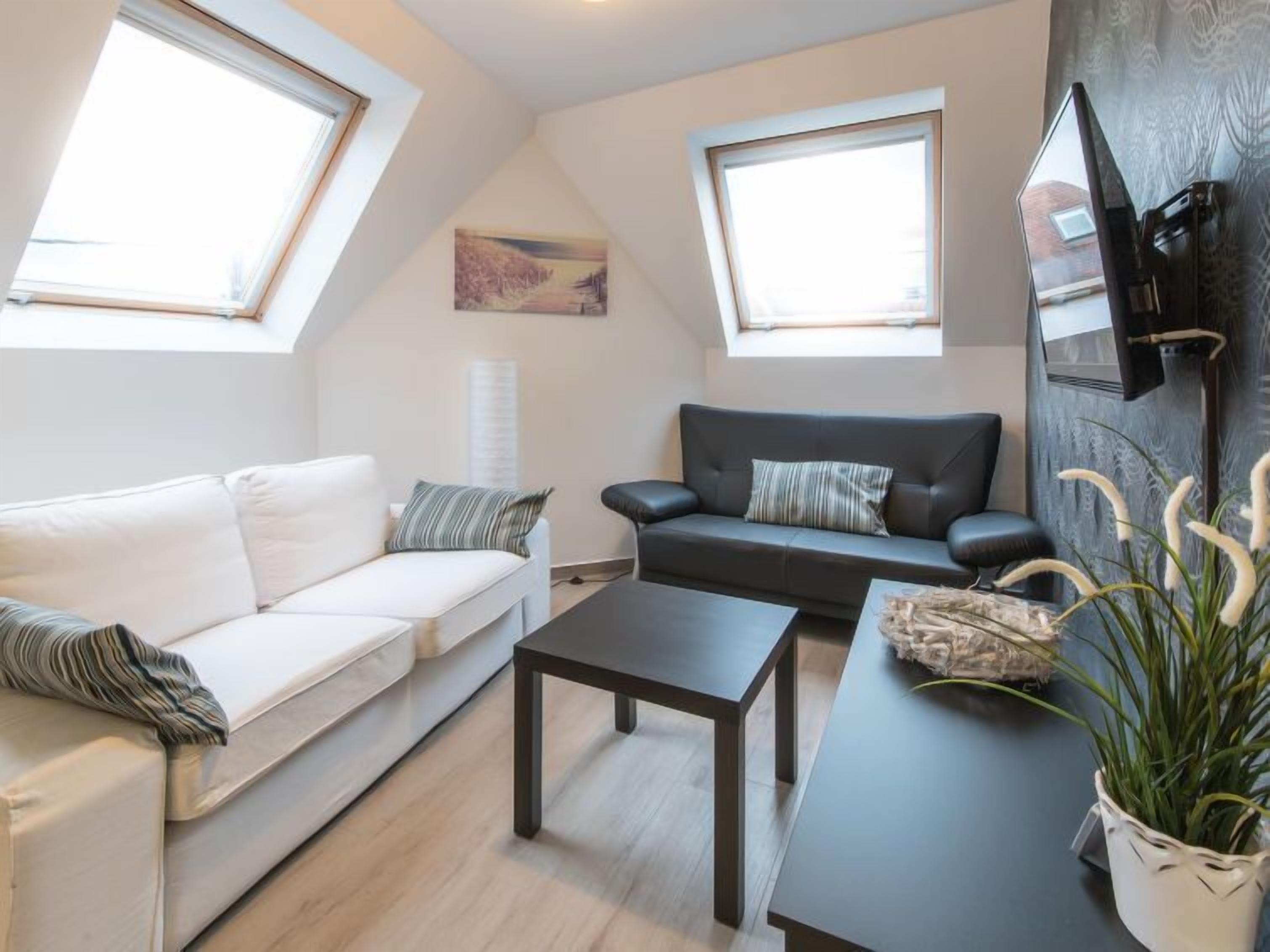 Un salon moderne avec un canapé blanc, un canapé foncé, une table basse et un téléviseur fixé au mur. Deux puits de lumière zénithaux permettent de profiter de la lumière naturelle.