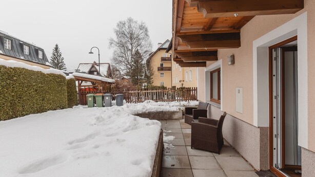 Un espacio exterior cubierto de nieve con un pérgola de madera, un área de descanso con mobiliario de mimbre y un edificio con balcón.