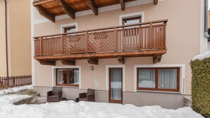 Un edificio de dos pisos con un balcón de madera, un área de estar al aire libre cubierta y el suelo cubierto de nieve.