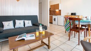 Ett modernt vardagsrum med en mörkgrå soffa, ett träbord och en randig stol. I bakgrunden syns ett kök med träskåp och en tvättmaskin.