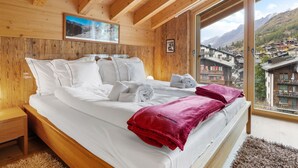 A wooden bedroom with a large bed, a view of a village through a window, and a painting on the wall. - Rütschi by Interhome (Zermatt)